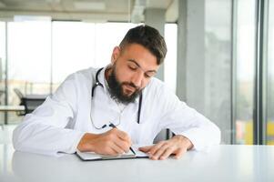 retrato de Arábica médico hermoso joven hombre en ropa de trabajo posando a moderno clínica, sentado a trabajo de Escritorio foto