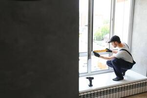 Workman in overalls installing or adjusting plastic windows in the living room at home photo