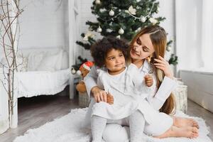 alegre Navidad y contento Días festivos alegre mamá y su linda hija a Navidad árbol. padre y pequeño niño teniendo divertido cerca Navidad árbol adentro. amoroso familia con regalos en habitación. foto