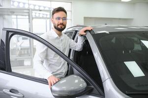 contento joven chico comprobación nuevo lujo auto, comprando automóvil a concesión centro. retrato de alegre milenario caucásico hombre examinando auto a sala de exposición Tienda foto