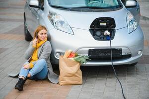 usando el teléfono inteligente mientras espera. mujer en la estación de carga de coches eléctricos durante el día. vehículo nuevo foto