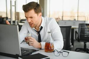 Young stressed handsome businessman working at desk in modern office shouting at laptop screen and being angry about financial situation, jealous of rival capabilities, unable to meet client needs photo