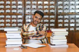 Portrait of cheerful male international Indian student with backpack, learning accessories standing near bookshelves at university library or book store during break between lessons. Education concept photo