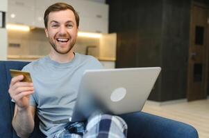 Happy Caucasian man buying things online, using smartphone, laptop and credit card, enjoying shopping in internet. Positive young guy purchasing goods on web, making remote payment photo