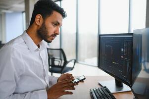 Side view of smart Indian crypto trader, investor, analyst broker, using laptop and smartphone analyzing digital cryptocurrency exchange, stock market charts, thinking of investing and funds risks. photo