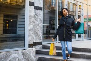 hermosa Rizado peludo afroamericano mujer en pie en un compras centro comercial con de colores compras pantalones en manos. foto