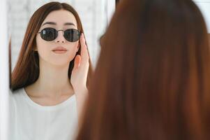 ella hecho su elección. hermosa joven mujer ajustando su nuevo Gafas de sol y sonriente mientras en pie en óptico almacenar. foto