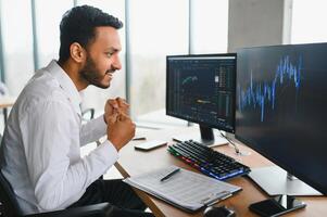 Side view of smart Indian crypto trader, investor, analyst broker, using laptop and smartphone analyzing digital cryptocurrency exchange, stock market charts, thinking of investing and funds risks. photo