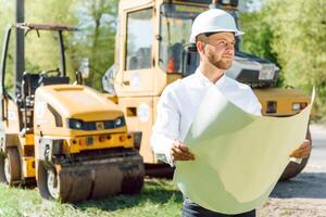 Smiling engineer with helmet standing in front of excavator on road construction site photo