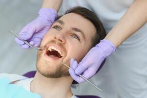 periodontal servicios. de cerca Disparo de sonriente hombre consiguiendo tratamiento en estomatológico clínica, dentista médico en guantes utilizando estéril dental herramientas para examinando dientes de masculino paciente foto