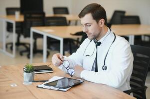 estresado masculino médico se sentó a su escritorio. medio adulto masculino médico trabajando largo horas. sobrecargado de trabajo médico en su oficina. no incluso doctores son eximir desde agotamiento . foto