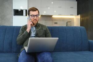 Cheerful busy young caucasian male manager sits on sofa with computer, calls by phone, talks with client, looks at free space in white room interior. Business, work remotely at home, ad and offer. photo