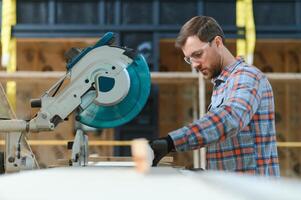 A carpenter works on woodworking the machine tool. Man collects furniture boxes. Saws furniture details with a circular saw. Process of sawing parts in parts. Against the background of the workshop. photo