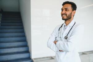 retrato de masculino indio médico vistiendo blanco Saco teniendo abierto puerta en clínica corredor como antecedentes foto