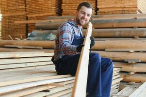 retrato de un hermoso trabajador elegir el mejor de madera tableros carpintero en pie siguiente a un grande apilar de madera barras en un depósito. foto