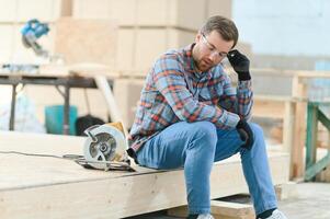 A carpenter works on woodworking the machine tool. Man collects furniture boxes. Saws furniture details with a circular saw. Process of sawing parts in parts. Against the background of the workshop. photo