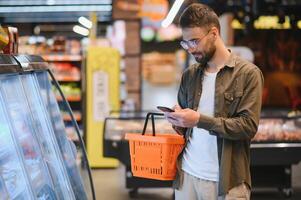 texto rápido durante las compras. apuesto joven sosteniendo un teléfono móvil y sonriendo mientras está de pie en una tienda de alimentos foto