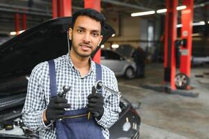 indio coche mecánico en pie y trabajando en Servicio estación. coche especialistas examinando el levantado coche. profesional reparadores vistiendo mecánico uniforme en azul color. foto