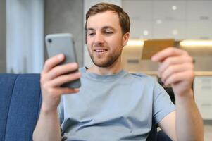 Happy Caucasian man buying things online, using smartphone, laptop and credit card, enjoying shopping in internet. Positive young guy purchasing goods on web, making remote payment photo