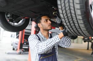 indio coche mecánico en pie y trabajando en Servicio estación. coche especialistas examinando el levantado coche. profesional reparadores vistiendo mecánico uniforme en azul color. foto