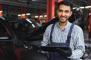 mecánico hombre mecánico gerente trabajador utilizando un ordenador portátil computadora comprobación coche en taller a auto coche reparar Servicio centro. ingeniero joven hombre mirando a inspección vehículo detalles debajo coche capucha. foto