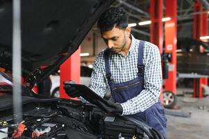 indio coche mecánico en pie y trabajando en Servicio estación. coche especialistas examinando el levantado coche. profesional reparadores vistiendo mecánico uniforme en azul color. foto