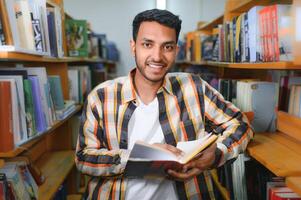 Portrait of cheerful male international Indian student with backpack, learning accessories standing near bookshelves at university library or book store during break between lessons. Education concept photo