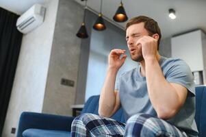 Feeling stressed. Frustrated handsome young man touching his head and keeping eyes closed while sitting on the couch at home photo