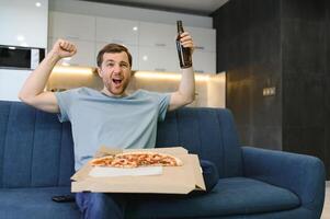 contento joven hombre Bebiendo cerveza y comiendo Pizza cuando acecho juego en televisión a hogar foto