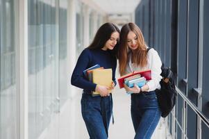 dos joven hembra estudiantes en pie con libros y pantalones en el pasillo Universidad Hablando cada otro. foto