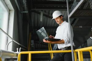 A young handsome employee of a modern factory in a white hard. In the background is a large shop for the production. photo