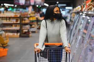 africano mujer vistiendo desechable médico mascarilla. compras en supermercado durante coronavirus pandemia brote. epidemia hora foto