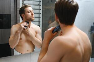 Reflection of young man in mirror shaving with electric shaver photo