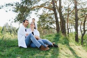 contento joven familia gasto hora al aire libre en un verano día tener divertido a hermosa parque en naturaleza mientras sentado en el verde césped. contento familia. foto