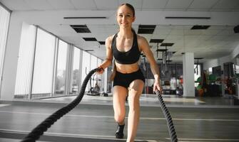 atlético hembra en un gimnasio ejercicios con batalla cuerdas durante su aptitud rutina de ejercicio Alta intensidad intervalo capacitación. ella es muscular y sudoroso foto