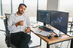 Side view of smart Indian crypto trader, investor, analyst broker, using laptop and smartphone analyzing digital cryptocurrency exchange, stock market charts, thinking of investing and funds risks. photo