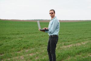 granjero en pie en joven trigo campo examinando cosecha y mirando a ordenador portátil. foto