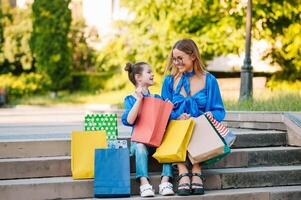 Beautiful mom and her cute little daughter are holding shopping bags, looking at camera and smiling while standing outdoors. Shopping concept. photo