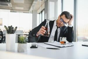 Fatigue young businessman sitting at workplace and holding whiskey bottle, drinking alcohol, bankruptcy concept. photo