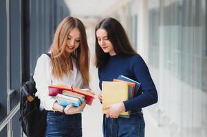 dos joven hembra estudiantes en pie con libros y pantalones en el pasillo Universidad Hablando cada otro. foto