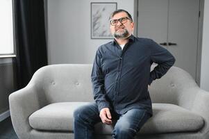 mayor hombre con blanco pelo y barba solo sentado en cama y sufrimiento desde bajo espalda dolor. antiguo edad, salud problema y personas concepto foto