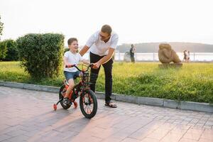 joven y contento padre enseña su joven hijo a paseo un bicicleta. el niño es contento . padre acecho hijo. del padre día foto