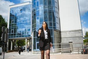 hermosa mujer yendo a trabajo con café caminando cerca oficina edificio. retrato de exitoso negocio mujer participación taza de caliente bebida en mano en su camino a trabajo en ciudad calle. alto resolución. foto