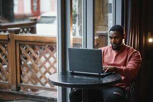 Portrait of happy african businessman using phone while working on laptop in restaurant. photo