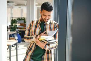 joven indio estudiante chico leyendo libro estudiando en Universidad biblioteca con estante para libros detrás. trabajando en asignación o proyecto foto