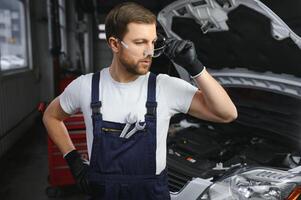 retrato de un joven hermosa coche mecánico en un coche taller, en el antecedentes de servicio. concepto reparar de máquinas, culpa diagnóstico, reparar especialista, técnico mantenimiento y a bordo computadora foto