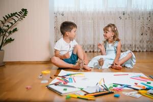 Kids drawing on floor on paper. Preschool boy and girl play on floor with educational toys - blocks, train, railroad, plane. Toys for preschool and kindergarten. Children at home or daycare. photo