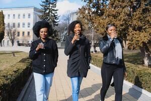 three beautiful and stylish dark-skinned girls with long hair standing in a city and drinking a coffee and use the phones photo