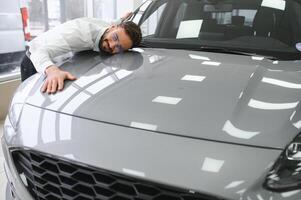 contento caucásico exitoso joven empresario abrazando abrazando su nuevo auto, sensación emocionado después comprando costoso auto a auto comerciante tienda almacenar. foto