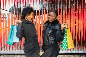 Portrait of young two african ladies friends standing over red wall and posing with shopping bags. photo